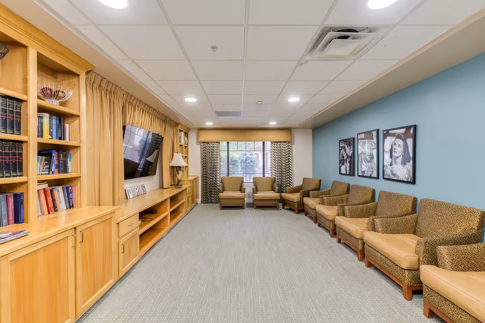 A spacious, well-lit common area with a row of cushioned armchairs along a blue wall adorned with three black and white framed portraits. Opposite the chairs is a wooden built-in shelving unit with books, decorative items, and a mounted flat-screen TV. A window with patterned curtains allows natural light into the room.