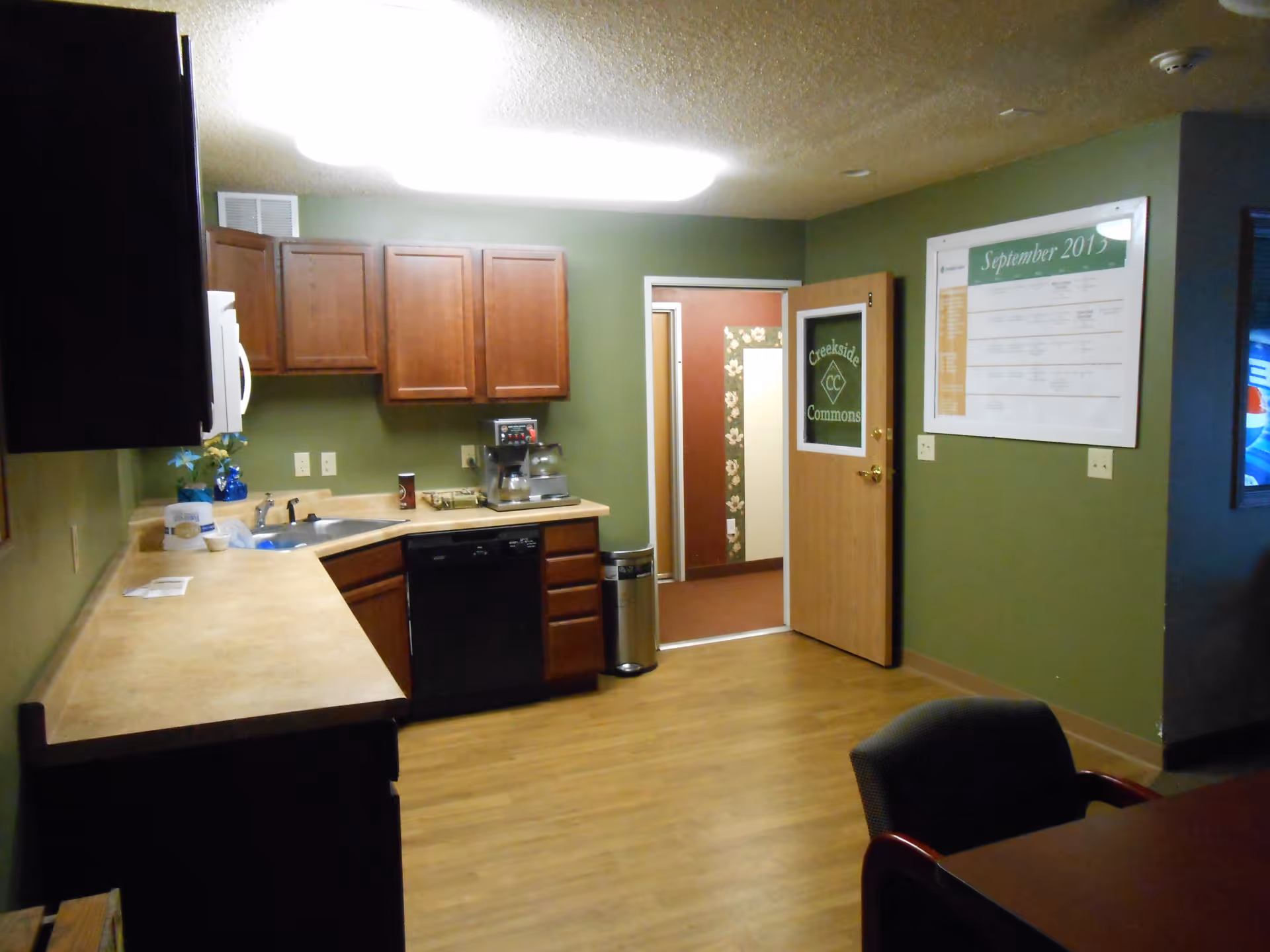 Interior view of a kitchen area with wooden cabinets, a countertop, a sink, a dishwasher, a coffee maker, and a trash can. The walls are painted green, and there is a door with a glass panel labeled 'Creekside Commons' leading to another room. A large calendar for September 2013 is mounted on the wall.