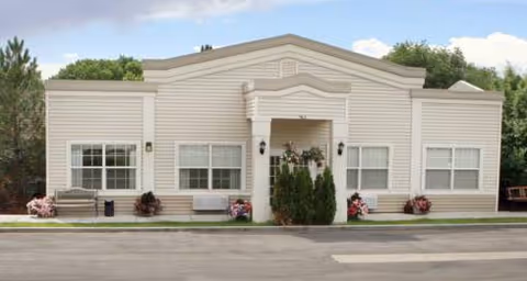 Single-story building with beige siding and a central entrance flanked by two windows on each side. There are small bushes and flower pots near the entrance and windows. The building is set against a backdrop of trees and a partly cloudy sky.
