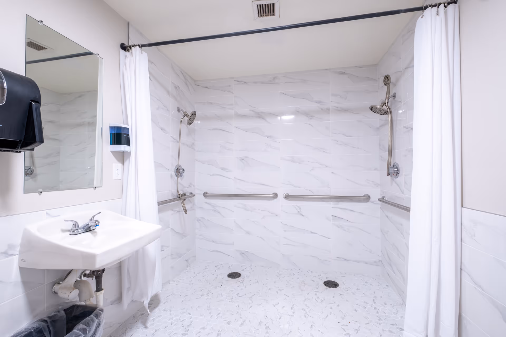 A clean, accessible bathroom with two shower heads mounted on opposite walls, white marble-patterned tiles, grab bars along the walls, a white sink with a mirror above it, a soap dispenser, and a white shower curtain.