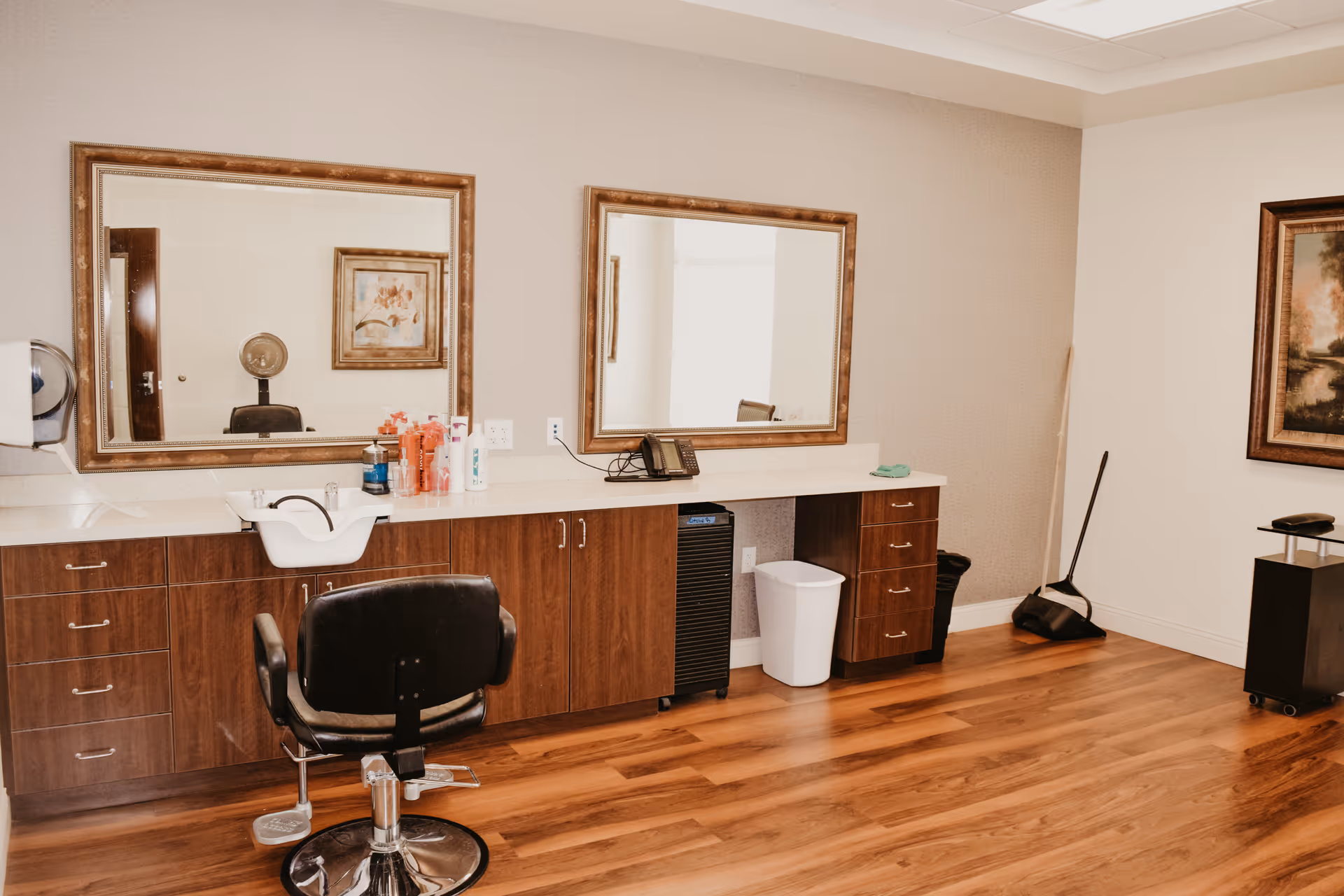 Interior of a salon or grooming area with a black salon chair in front of a countertop with a white sink, various hair care products, two large framed mirrors on the wall, wooden cabinets below the countertop, a white trash bin, a broom and dustpan in the corner, and framed artwork on the walls.