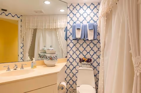 A bathroom with a white countertop and sink, a large mirror above the sink, and a toilet next to the counter. The walls have a blue and white patterned wallpaper. There are two blue towels hanging on a rack above the toilet, and a decorative ceramic container on the countertop. A shower with a white lace curtain is visible on the right side.