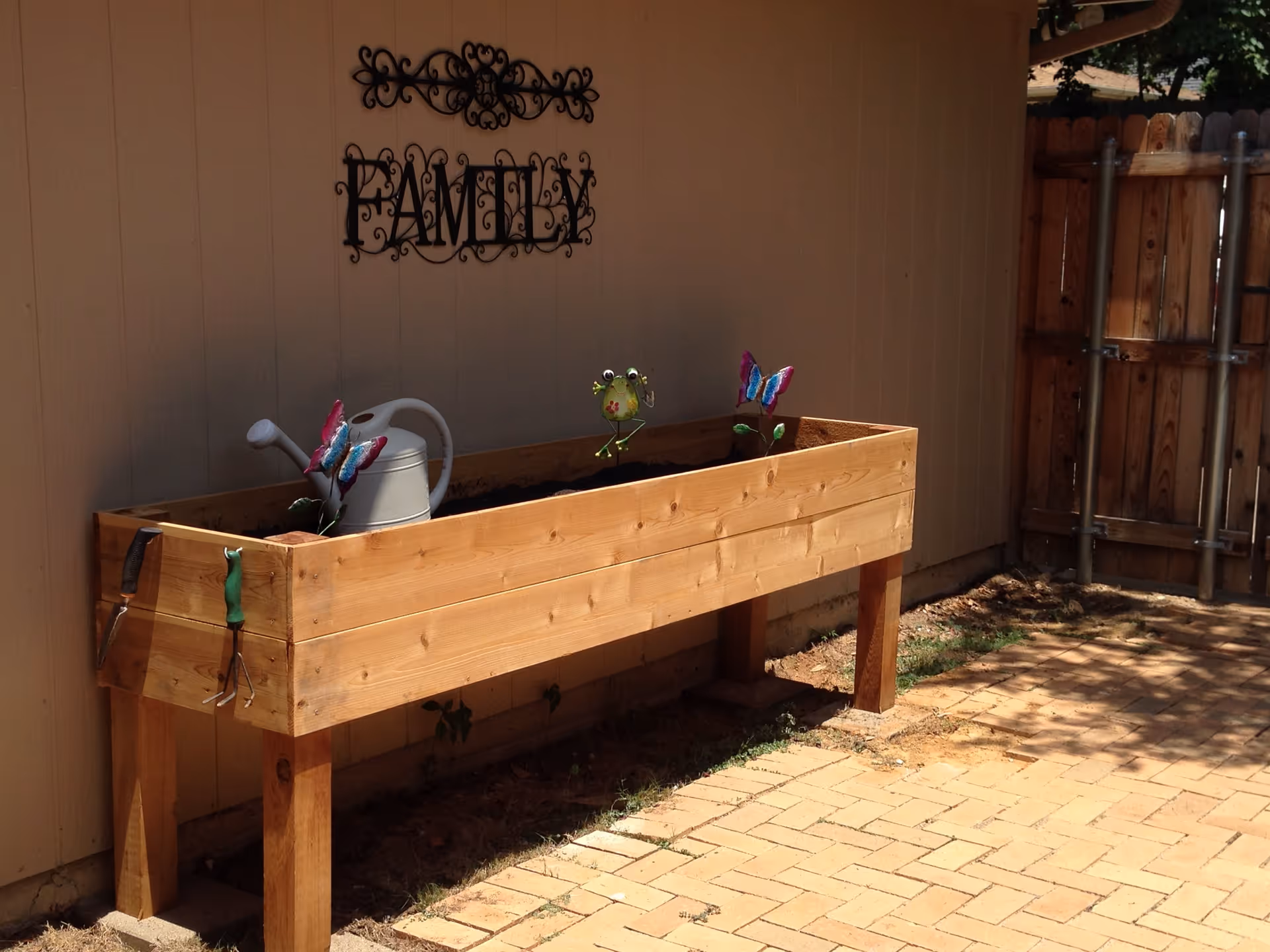 A wooden raised garden bed filled with soil, decorated with two colorful butterfly ornaments and a small frog ornament. A white watering can is placed inside the garden bed. Gardening tools hang on the side of the bed. The garden bed is positioned against a beige wall with a decorative black metal wall art that reads 'FAMILY'. The area is paved with brick tiles and there is a wooden fence in the background.