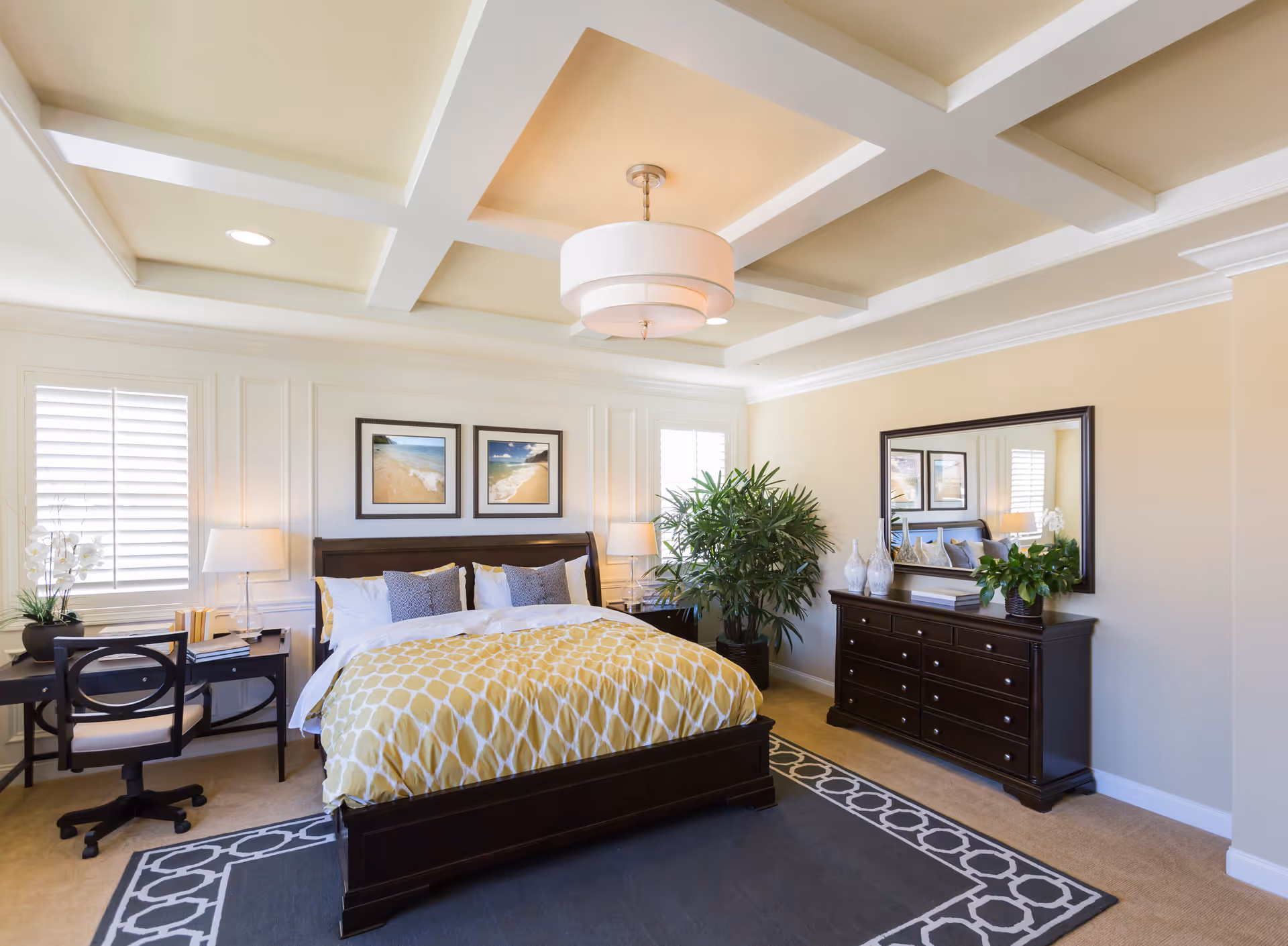 A bright and spacious bedroom with a large bed featuring a yellow and white patterned comforter and multiple pillows. The room has two windows with white shutters, a dark wooden dresser with a large mirror above it, and a desk with a chair and lamp. There are two framed beach-themed pictures above the bed and a large green plant next to the dresser. The ceiling has a coffered design with a modern light fixture.