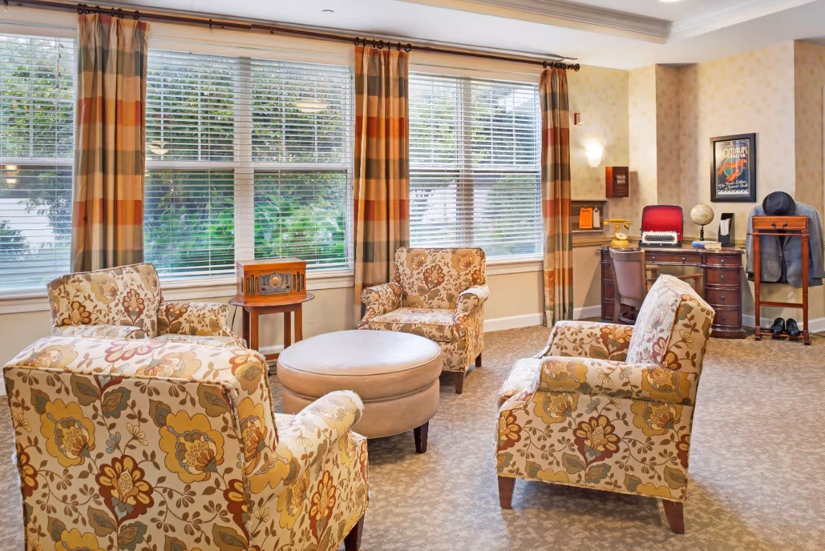 Sunny common room with four floral armchairs around a round ottoman, large windows, and a desk in the corner.