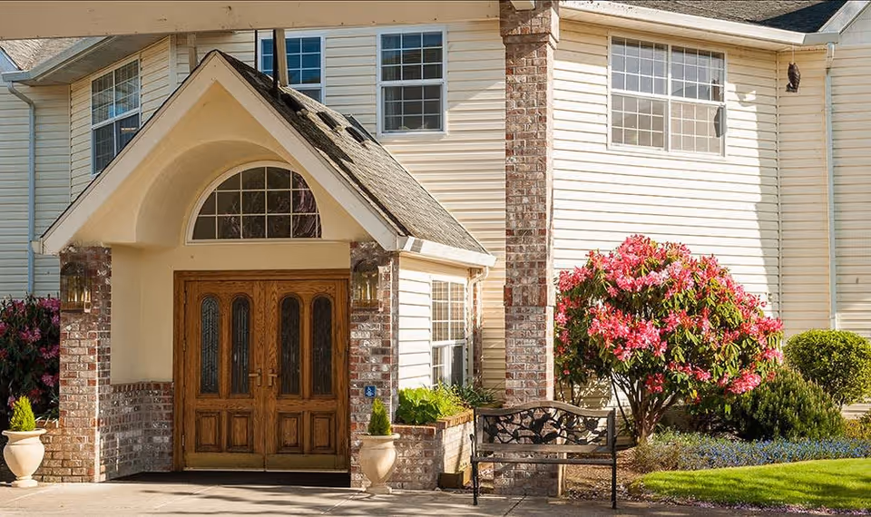 Entrance of a senior living facility with double wooden doors under a peaked roof supported by brick columns. There are large windows above and beside the doors. A black metal bench sits near a blooming pink rhododendron bush and other greenery on a well-maintained lawn.