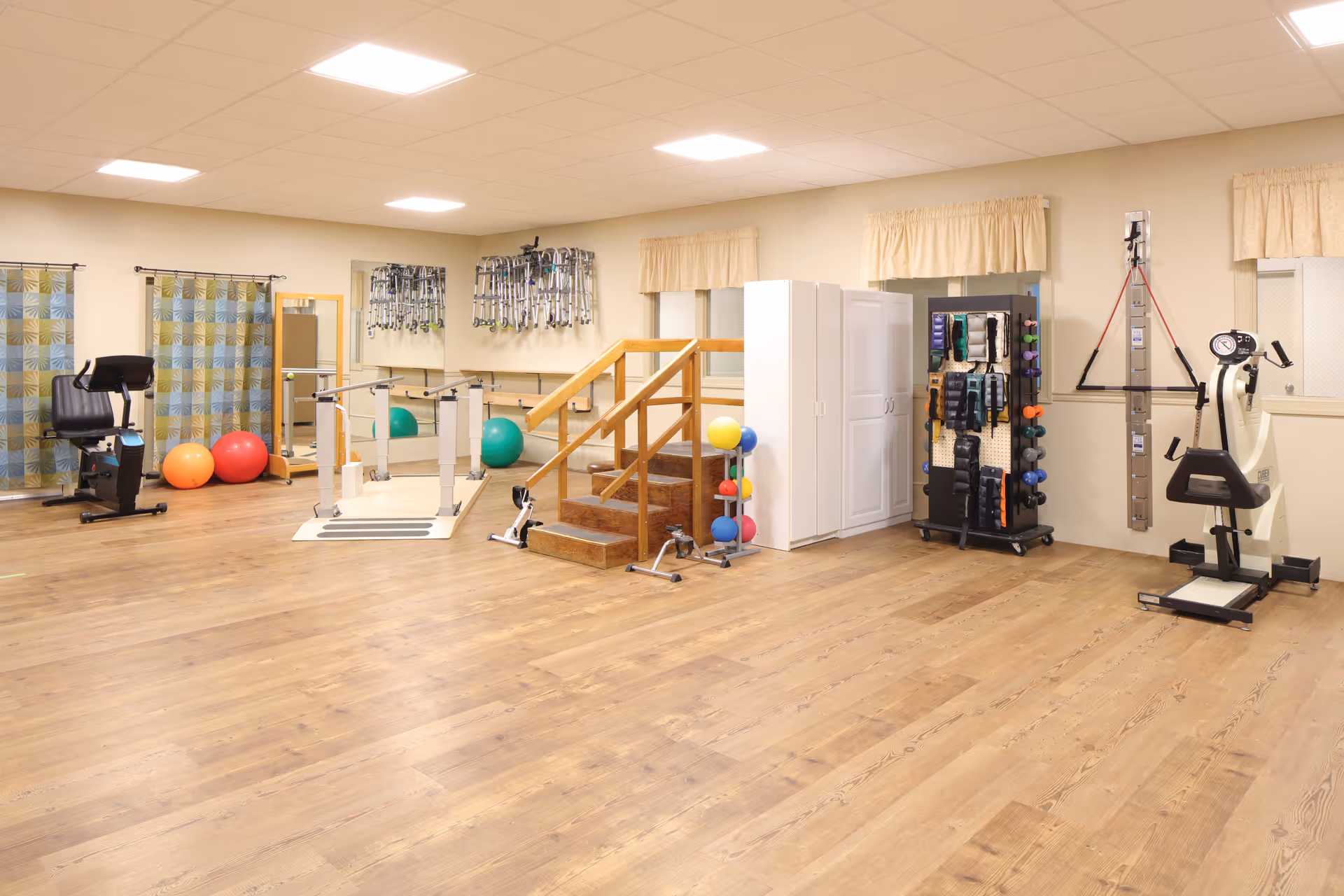 A spacious rehabilitation room with wooden flooring, exercise equipment including stationary bikes, parallel bars, therapy stairs, exercise balls, and various physical therapy tools organized on racks and shelves. The room has beige walls, windows with curtains, and bright ceiling lights.