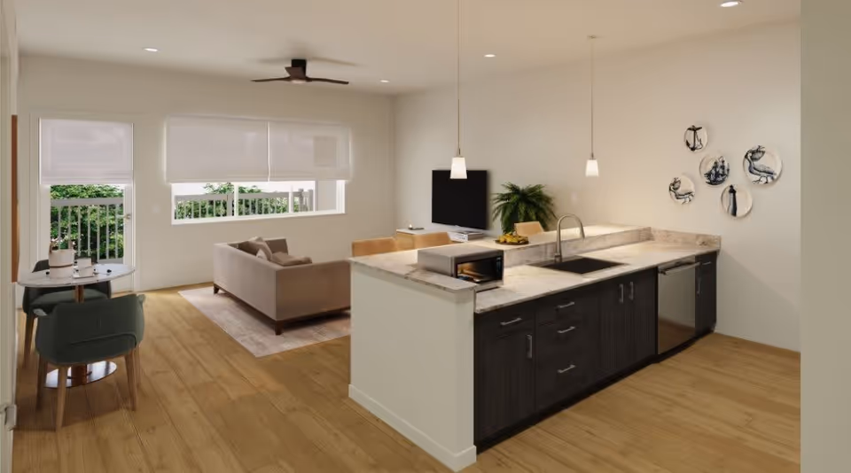A modern open-concept living space featuring a kitchen with dark cabinets, a marble countertop, a sink, and a microwave. Adjacent to the kitchen is a living room area with a beige sofa, a TV mounted on the wall, and a small dining table with two green chairs near a window. The room has light wood flooring, white walls, and decorative plates with nautical designs on one wall. Large windows with white blinds allow natural light to enter the space.