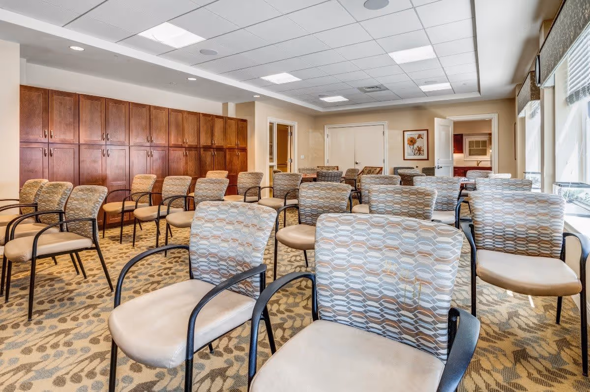 A well-lit room with multiple rows of cushioned chairs arranged facing forward, featuring patterned upholstery and beige seats. The room has a carpet with a leaf pattern, wooden cabinets along one wall, and large windows with blinds letting in natural light. There are framed pictures on the walls and a door leading to another room.