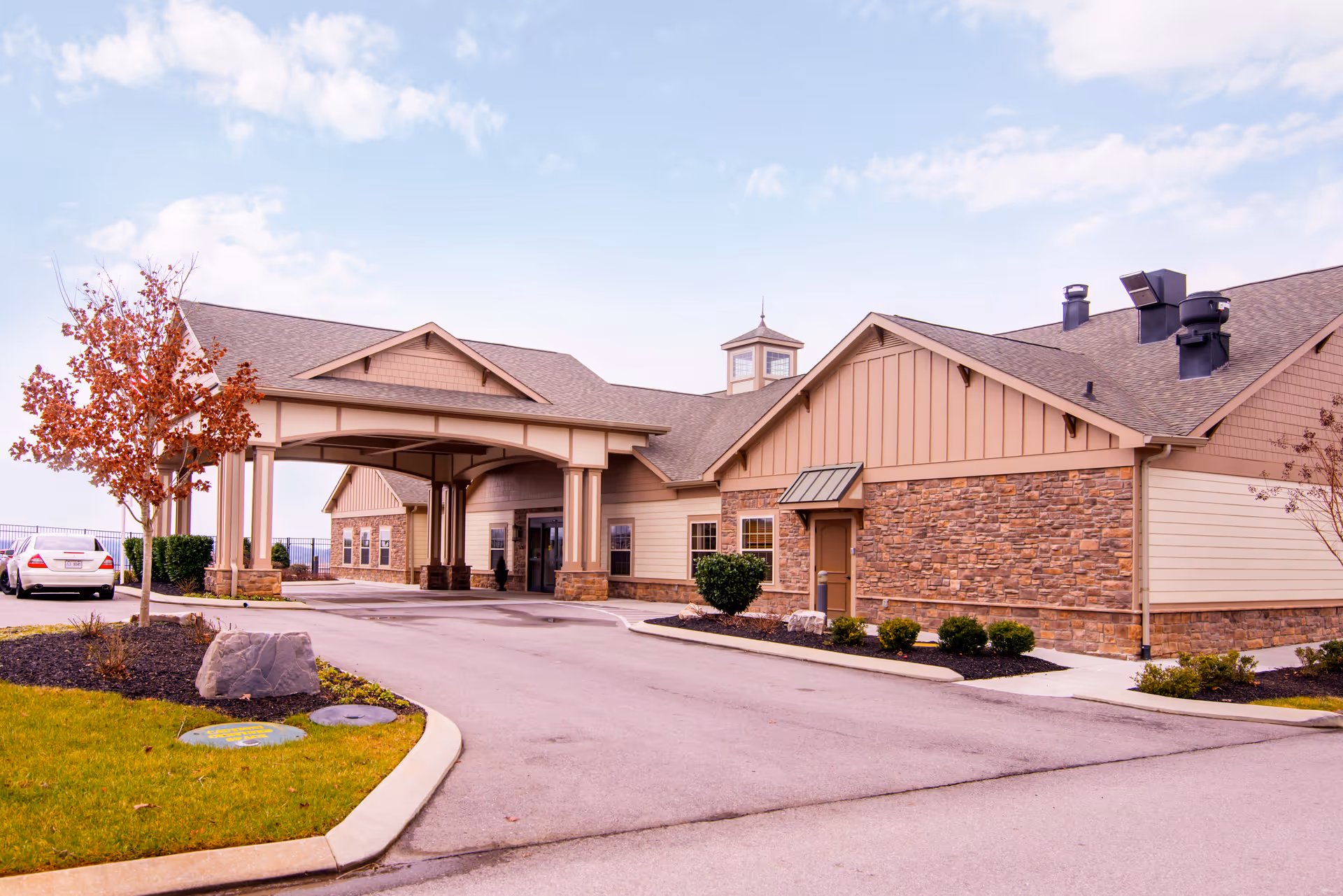 Front exterior of a single-story senior living facility with a covered drive-through entrance, stone and siding facade, and landscaped driveway.