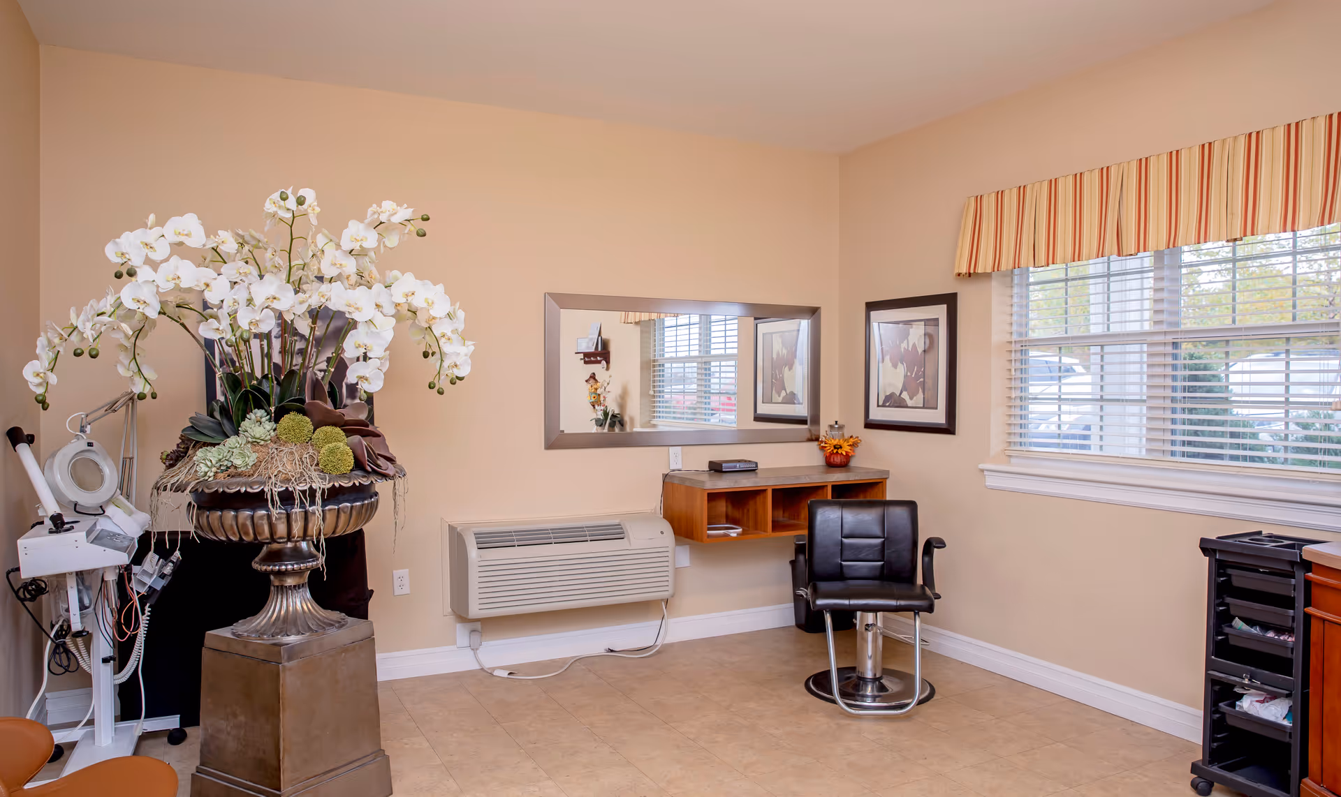 Bright salon-style room with a styling chair, wall mirror, large floral arrangement, and a window with blinds.