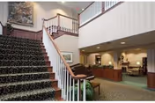 Lobby of a senior living community with a carpeted staircase, reception desk, and seating area under a balcony.