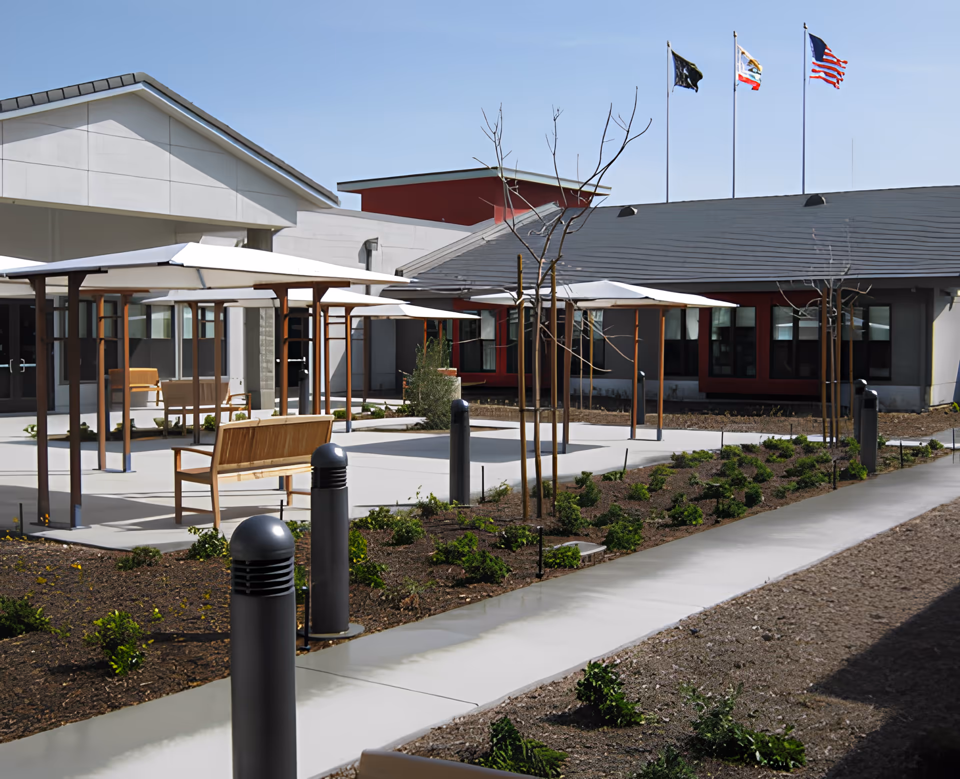 Sunlit courtyard with benches, shaded pergolas, young trees and walkways in front of a single-story building with flags flying above.