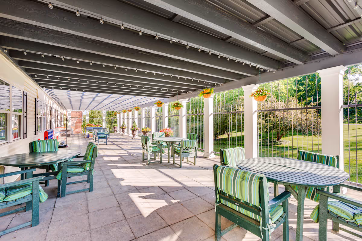Covered outdoor patio area with multiple round tables and green cushioned chairs. Hanging flower pots are suspended from the ceiling beams, and there is a metal railing along the side overlooking a grassy area with trees.