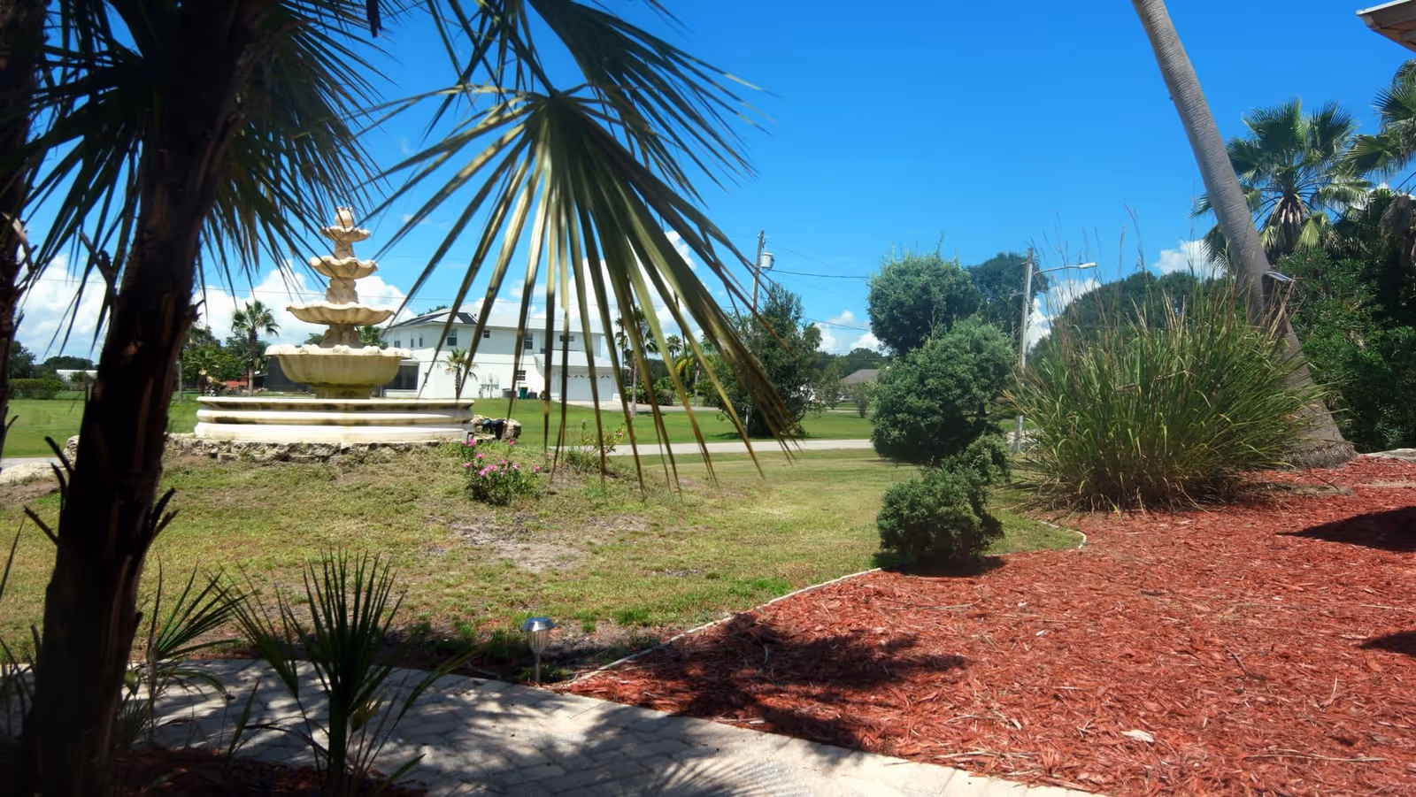 Outdoor garden area with a multi-tiered stone fountain, palm trees, trimmed bushes, and a mulched flower bed under a clear blue sky.