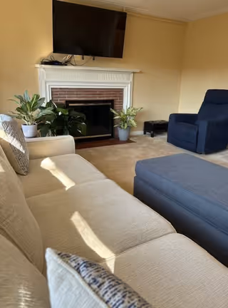 A cozy living room with a beige sofa and patterned cushions in the foreground, a blue armchair, a large blue ottoman, a white fireplace with a brick interior, a flat-screen TV mounted above the fireplace, and two potted plants on either side of the fireplace.