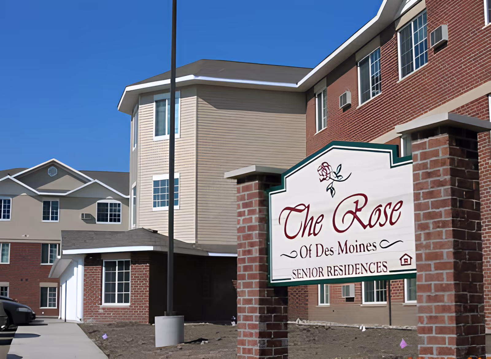 Exterior view of The Rose of Des Moines Senior Residences building with a clear blue sky. The building has a combination of beige siding and red brick walls with multiple windows. In the foreground, there is a sign with a rose graphic that reads 'The Rose Of Des Moines Senior Residences.'