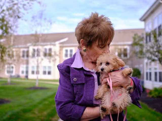 An elderly woman wearing a purple jacket is holding and kissing a small fluffy dog with a pink leash in an outdoor garden area of a senior living facility.