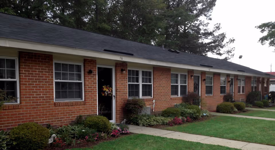 Single-story red brick apartment building with multiple front doors and windows and a landscaped lawn.