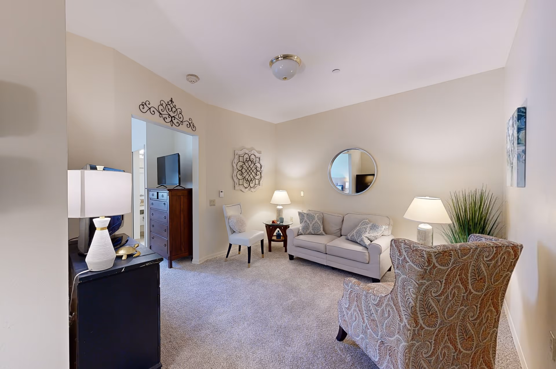 A cozy living room with a beige sofa adorned with patterned cushions, a patterned armchair, a white chair, two side tables with lamps, a round mirror on the wall, decorative wall art, a TV on a dresser visible through an open doorway, and a carpeted floor.
