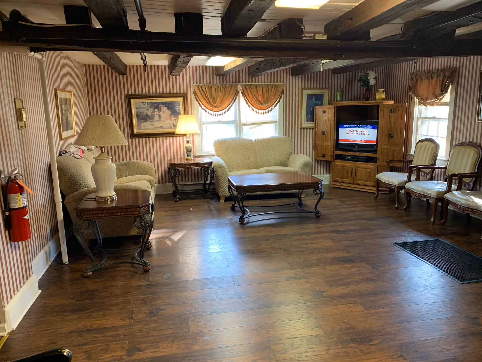 A cozy living room with wooden floors and exposed wooden ceiling beams. The room features a beige loveseat, a matching armchair, a wooden coffee table, and a wooden TV cabinet with a television displaying information about 2020 Medicare Advantage Plans. There are three wooden chairs with white cushions along the right wall, striped wallpaper, framed artwork, two windows with orange valances, and a table lamp on a side table. A fire extinguisher is mounted on the left wall.