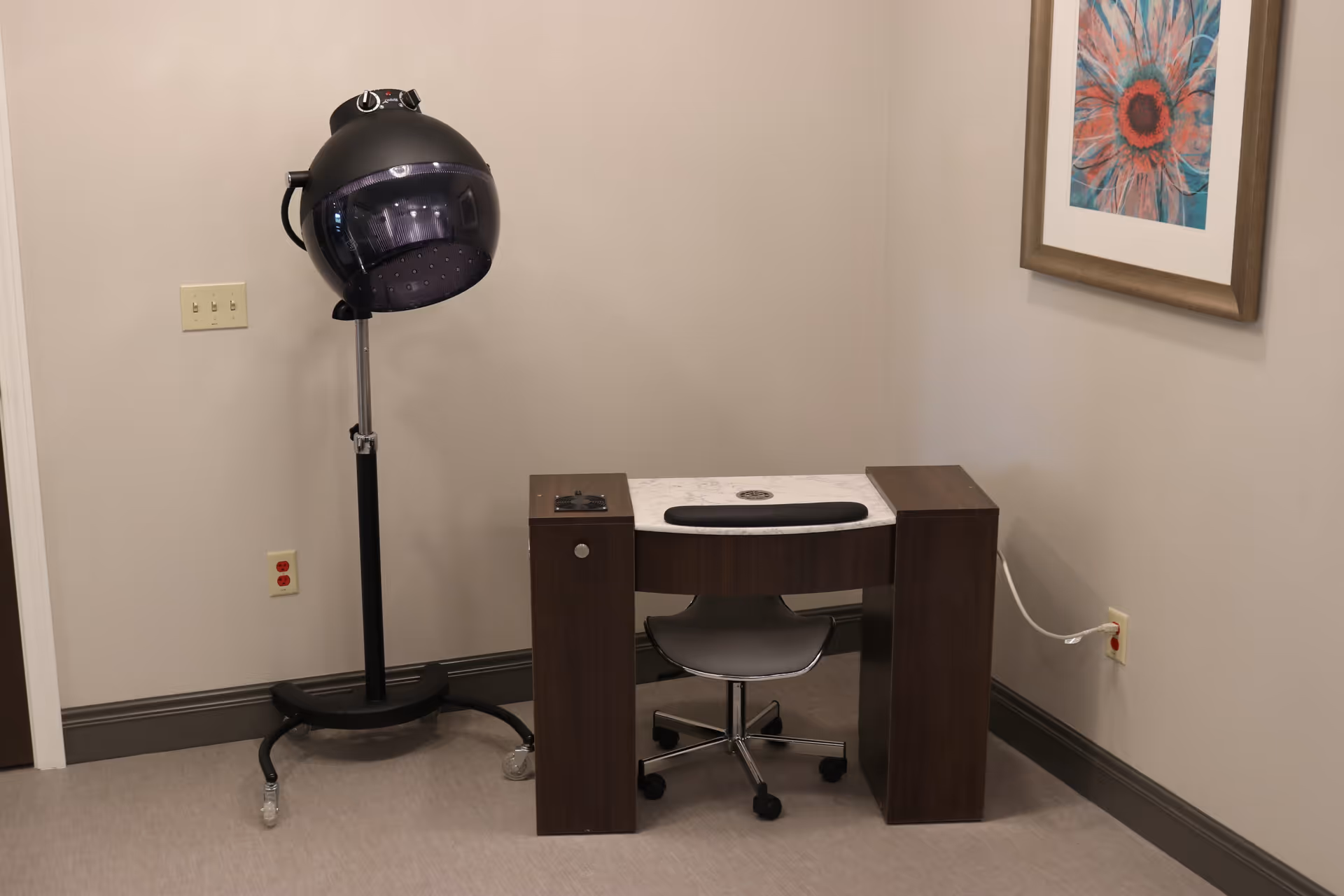 A small room with a hair drying station featuring a black hooded hair dryer on a stand with wheels, a dark wooden manicure table with a white top, a gray swivel chair, and a colorful framed flower artwork on the wall.