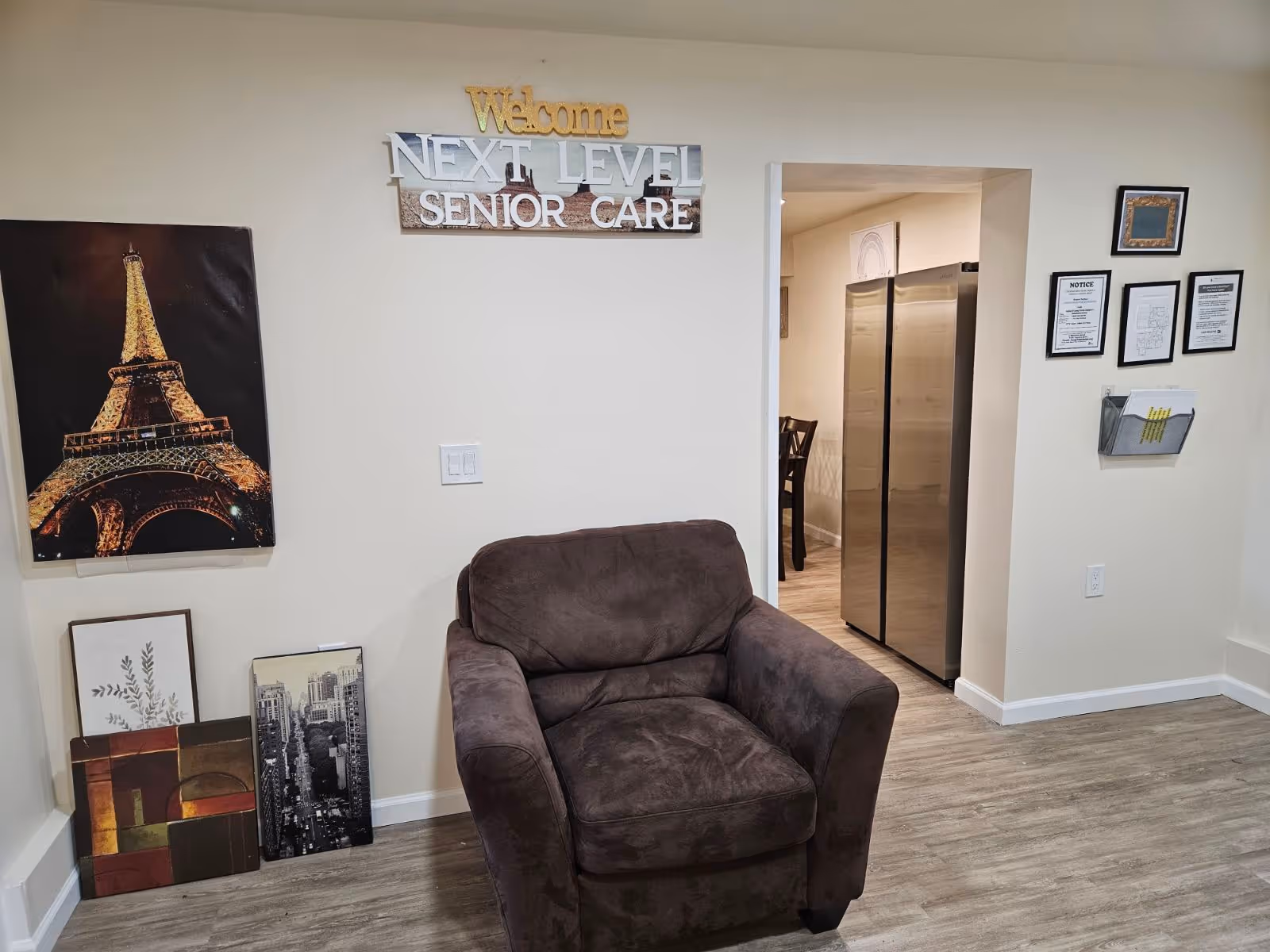A cozy interior seating area with a brown armchair beneath a 'Welcome Next Level Senior Care' sign and wall art, with a view into a kitchen/dining area.