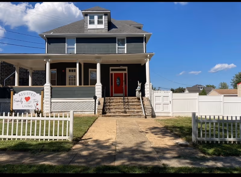 Exterior front view of a two-story assisted living facility named Family Ties Assisted Living. The building has a dark gray exterior with white trim, a red front door, a small porch with white columns, and a white picket fence surrounding the front yard. There is a sign on the left side of the yard that reads 'Assisted Family Ties Living'. The sky is clear with a few clouds.