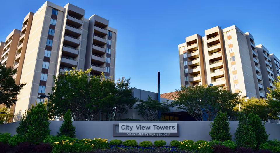 Exterior view of City View Towers Senior Apartments, showing two multi-story buildings with balconies surrounded by trees and landscaped greenery under a clear blue sky.