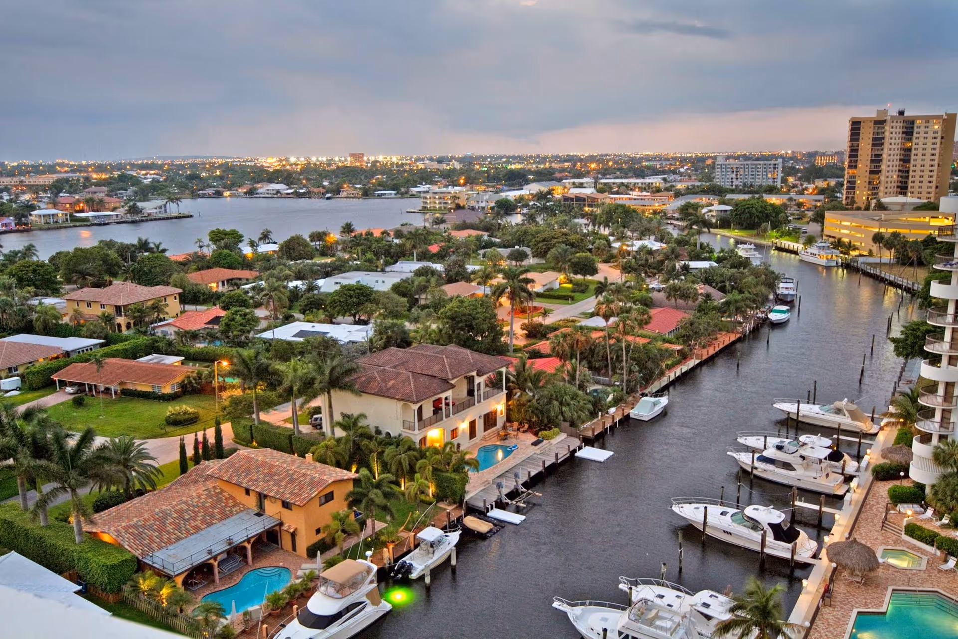 Aerial view of a waterfront residential neighborhood with houses, palm trees, and boats docked along a canal. The scene includes a mix of greenery, water, and buildings with city lights visible in the distance under a cloudy sky.