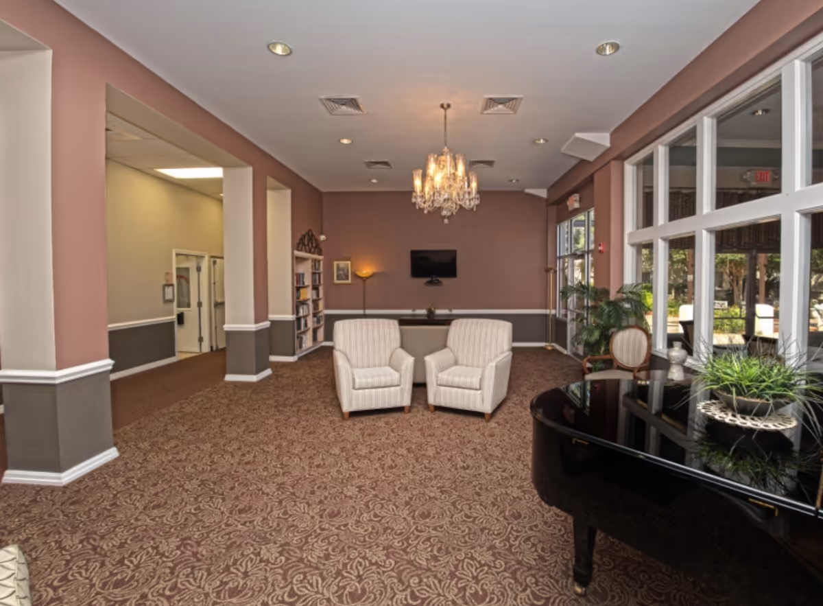 A spacious lounge area with patterned carpet, two beige armchairs facing a wall-mounted TV, a chandelier hanging from the ceiling, a bookshelf, a floor lamp, large windows letting in natural light, and a black grand piano with a plant on top.
