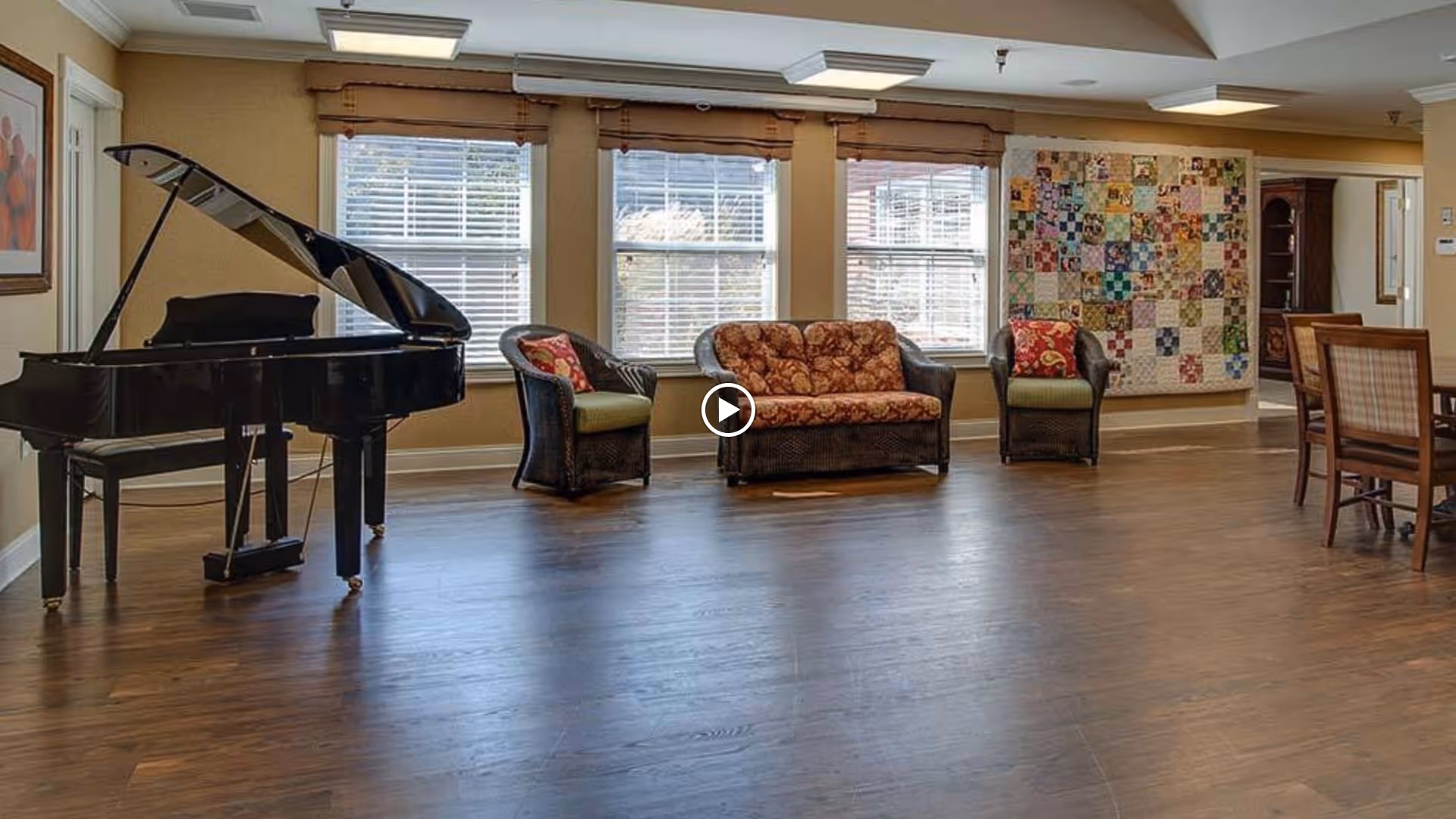A spacious senior living common area with a grand piano on the left, three wicker chairs with cushions arranged near large windows, and a colorful patchwork quilt hanging on the wall. There is also a wooden dining table with chairs on the right side of the room.