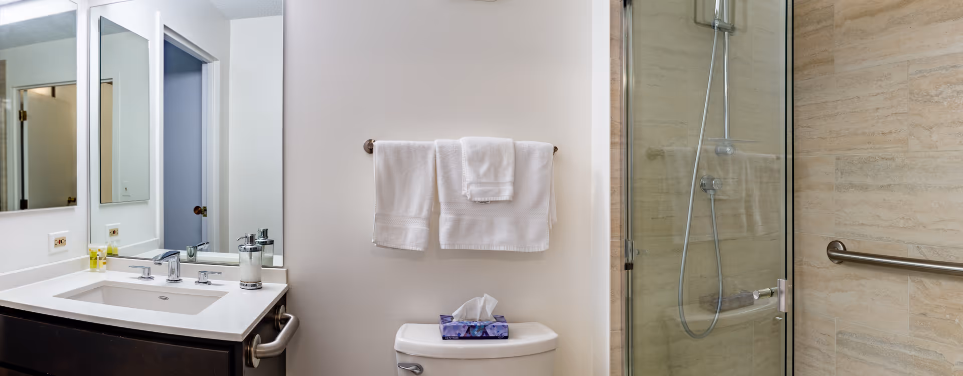 A clean and modern bathroom featuring a white sink with a dark wooden cabinet, a large mirror above the sink, a toilet with a tissue box on top, white towels hanging on a towel rack, and a glass-enclosed shower with beige tiled walls and a metal grab bar.