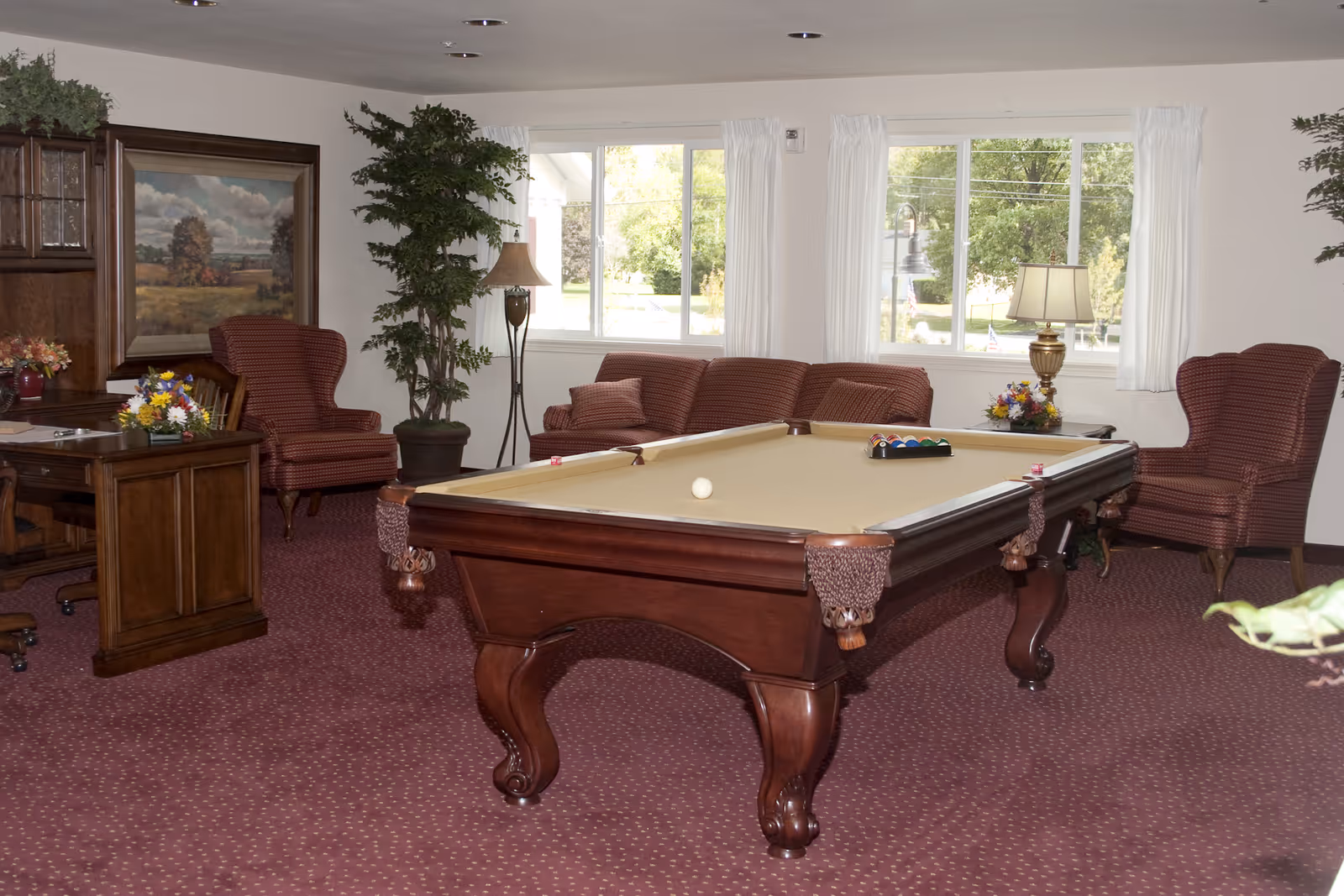 A senior living facility common room with a pool table in the center, surrounded by upholstered armchairs and a sofa. The room has large windows with white curtains letting in natural light, a wooden desk with flowers, a floor lamp, and framed landscape artwork on the wall.