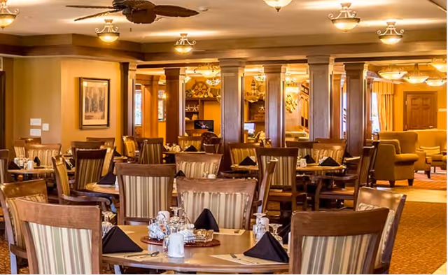 Warm, elegantly furnished dining room with round tables set with place settings and upholstered chairs in a senior living facility.