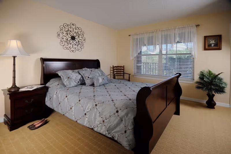 A bright bedroom with a dark wooden sleigh bed, nightstand and lamp, window with blinds and lace curtains, and a potted plant.
