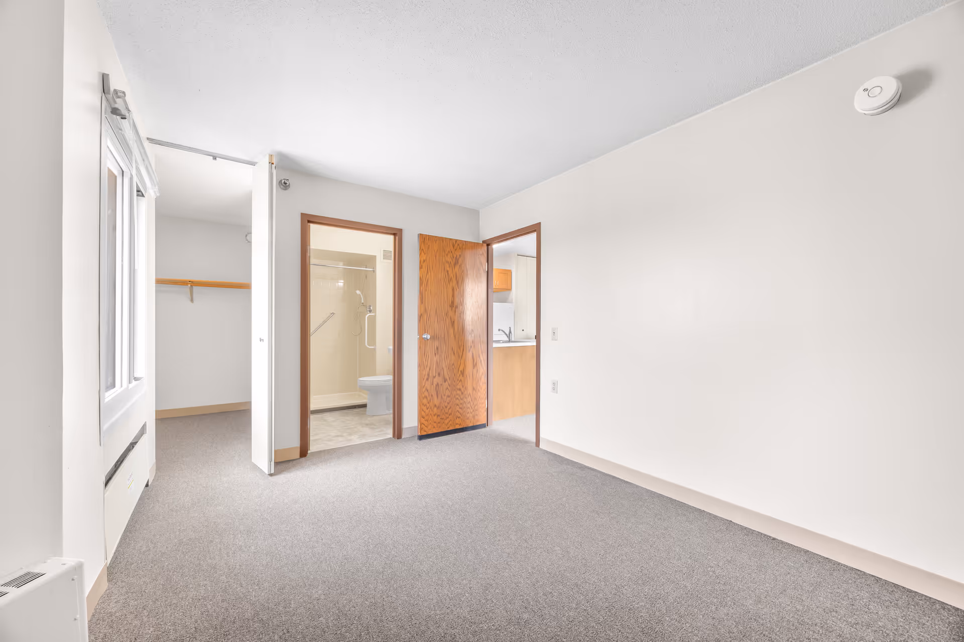 Empty room with light gray carpet and white walls. There is a window on the left side, an open closet with a wooden shelf, a doorway leading to a bathroom with a shower and toilet, and another doorway leading to a kitchen area with wooden cabinets and a sink.