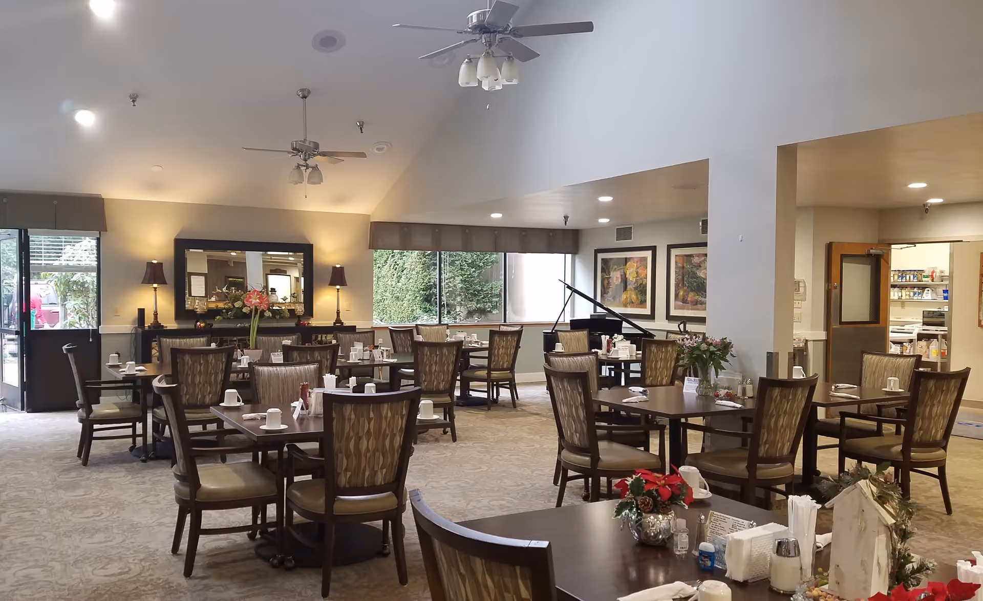 A spacious dining room with multiple wooden tables and chairs arranged neatly. Each table is set with cups, napkins, and condiments. The room features large windows with views of greenery outside, ceiling fans with lights, and decorative lamps on a sideboard with a large mirror above it. There is a grand piano in the corner and framed artwork on the walls. The room has a warm and inviting atmosphere.