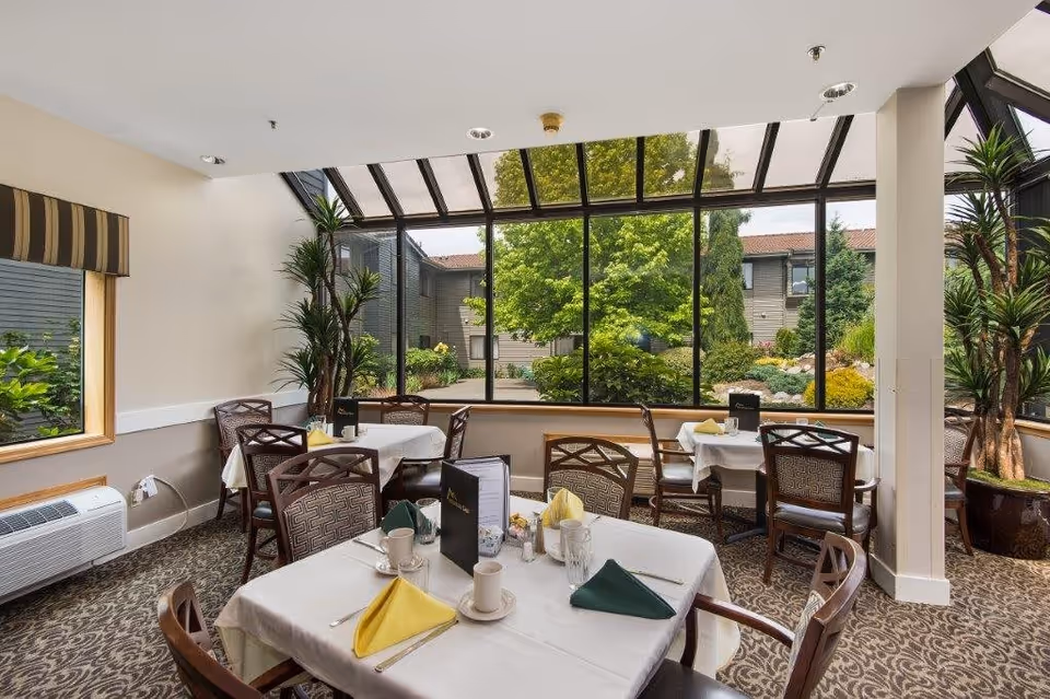 A bright dining room with several tables covered in white tablecloths, each set with cups, glasses, utensils, and folded napkins in yellow and green. The room features large windows and a glass ceiling that provide a clear view of a lush garden with trees and shrubs outside. There are patterned carpet floors and potted plants inside the room.