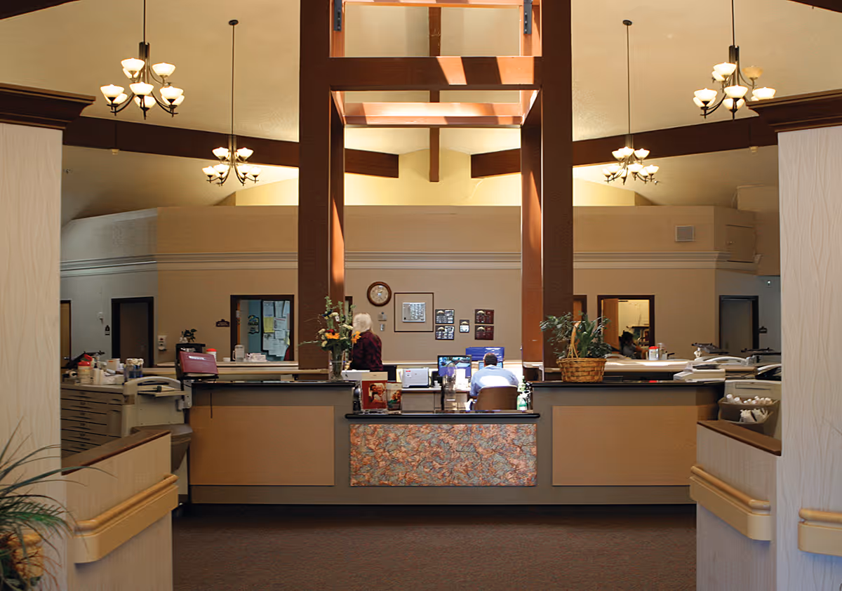 Reception area inside Sandstone North Park facility with a front desk, two people working behind the desk, decorative plants, and ceiling chandeliers.