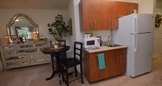 A small kitchenette area with wooden cabinets, a white refrigerator, a microwave on the counter, and a sink. Next to the kitchenette is a small round black table with two black chairs. In the background, there is a large decorative mirror above a dresser with lamps and plants, and a tall potted plant beside the table.