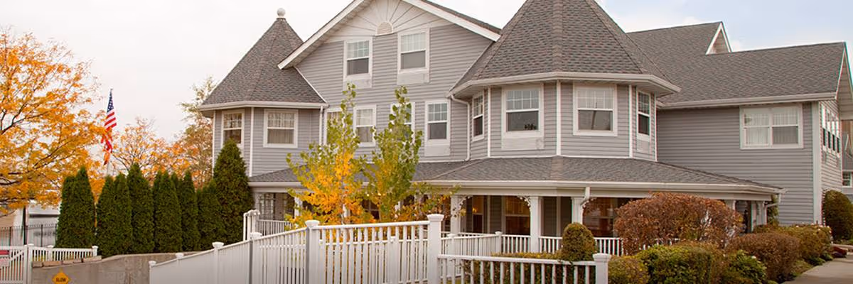 Exterior view of a large, gray senior living facility with multiple peaked roofs and white trim. The building is surrounded by a white fence, green shrubs, and trees with autumn foliage. An American flag is visible on the left side near the building.