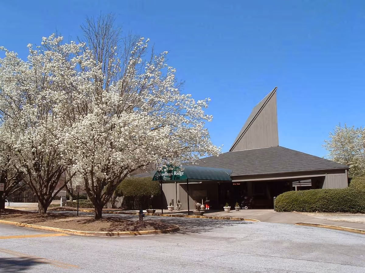 Exterior view of Woodland Ridge Assisted Living facility with a unique peaked roof design, surrounded by blooming white flowering trees and trimmed bushes under a clear blue sky.