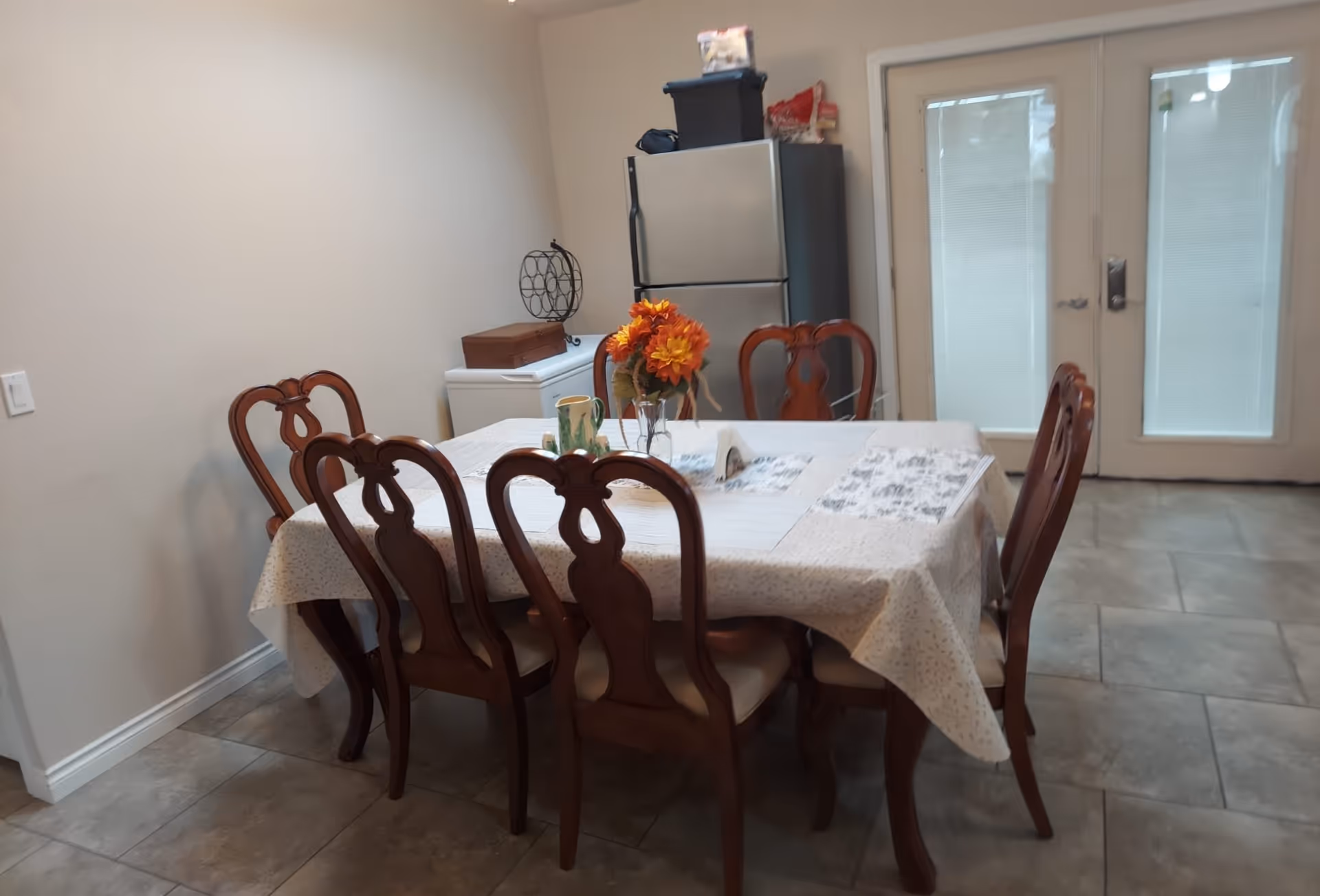 Dining room with a rectangular table covered by a tablecloth and six wooden chairs, a vase of flowers on the table, and a refrigerator and double glass doors in the background.