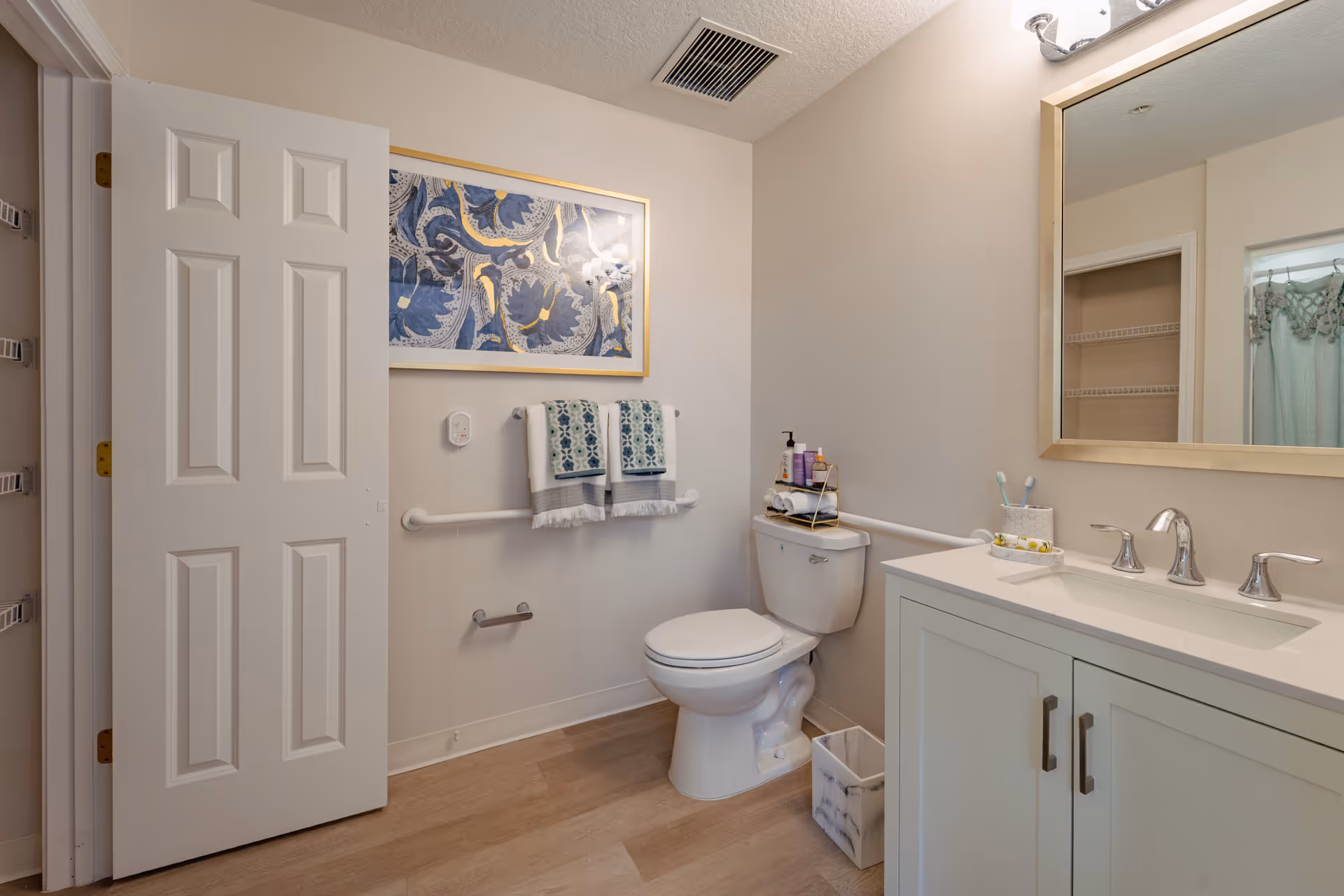 A clean and well-lit bathroom featuring a white toilet with a small rack holding toiletries on top, a white vanity with a sink and silver faucet, a large mirror above the sink, a towel rack with two patterned towels, a framed blue and gold artwork on the wall, and a partially open door revealing a shower curtain in the adjacent room.