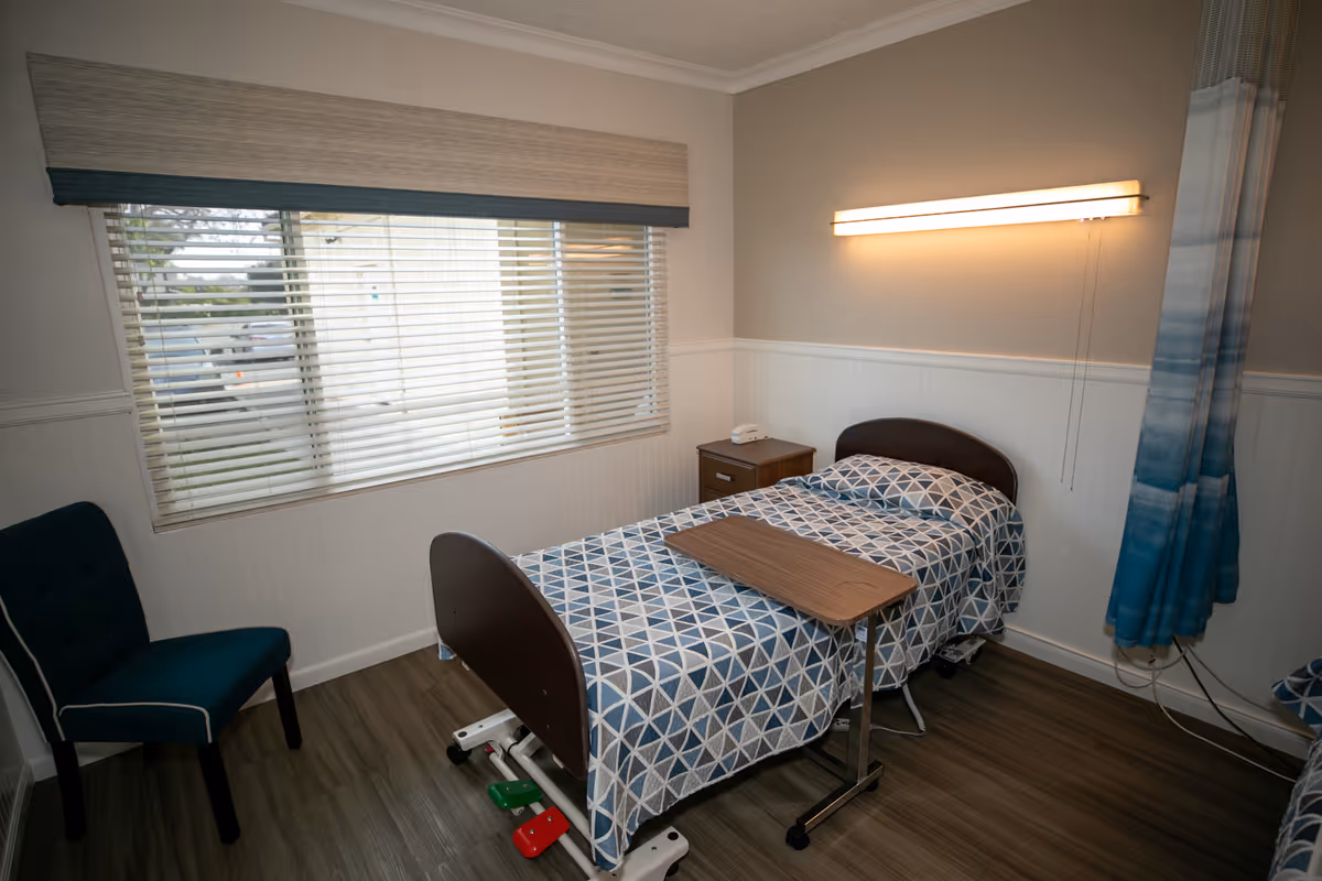 A small bedroom in a healthcare facility with a single bed covered in a blue and white geometric patterned blanket. There is a wooden bedside table with a white telephone on it, a blue upholstered chair, a window with blinds, and a wall-mounted light above the bed. A blue privacy curtain is partially visible on the right side.