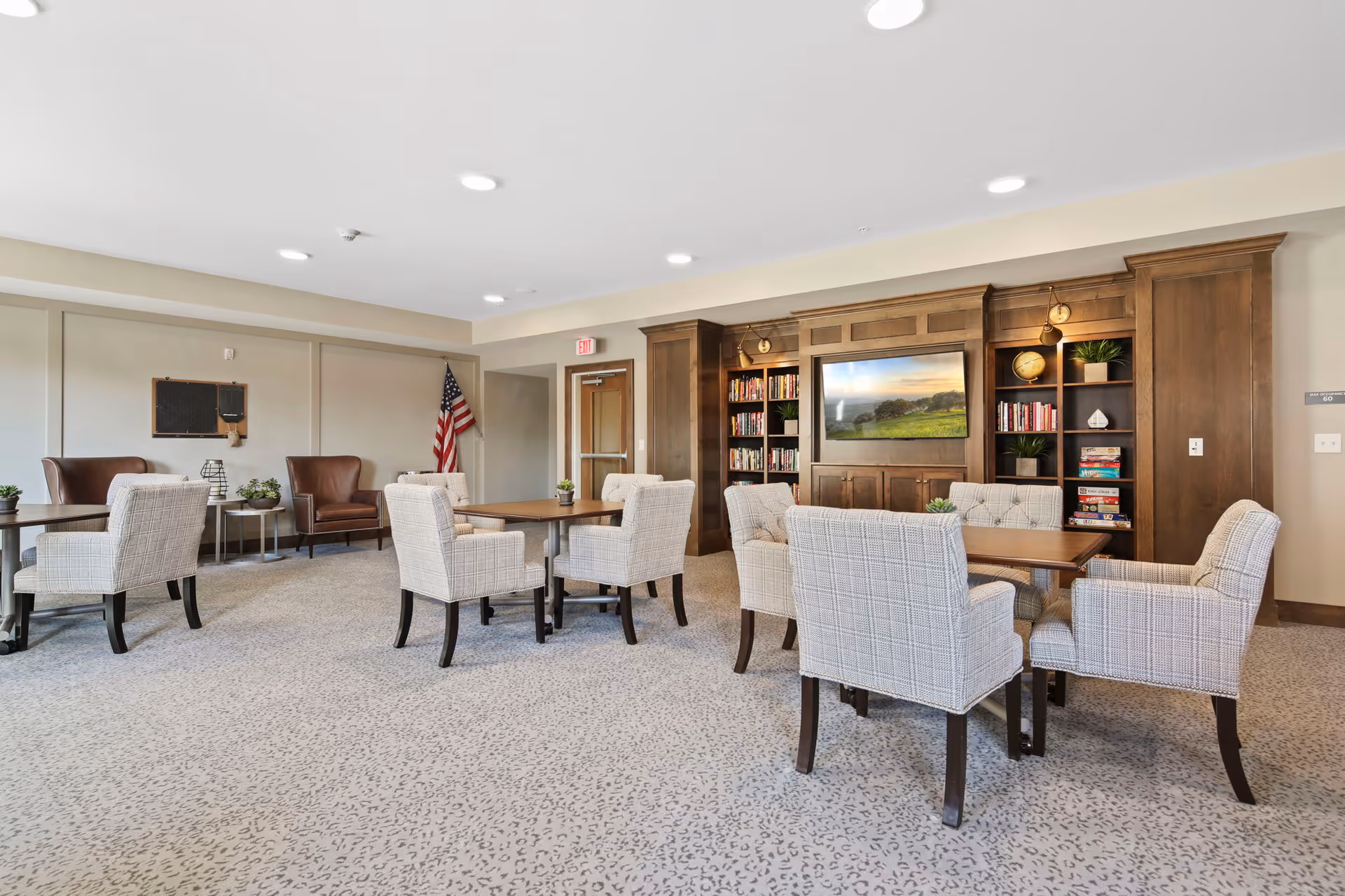 A bright and spacious common area with several tables surrounded by upholstered chairs. The room features a built-in wooden bookshelf with books, plants, and board games, and a large TV mounted in the center. There are two leather armchairs near the back wall, an American flag in the corner, and neutral-colored carpet and walls.