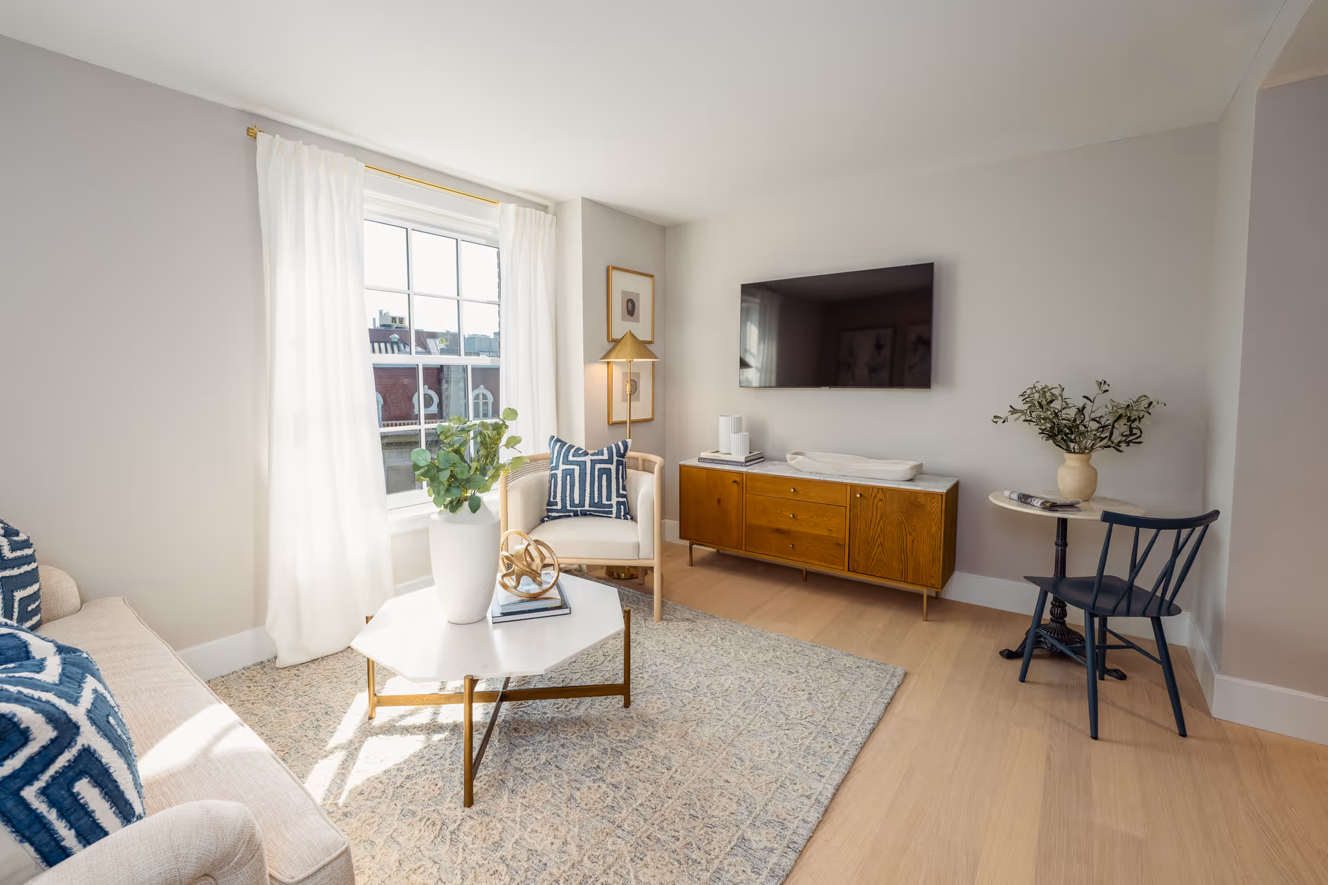 A bright and modern living room with light wood flooring, a large window with white curtains, a beige sofa with blue and white patterned pillows, a white armchair with a matching pillow, a white hexagonal coffee table with a white vase and decorative items, a wooden media console with a flat-screen TV mounted above it, and a small round side table with a vase of greenery and a black chair.