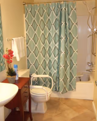 A bathroom with a white toilet equipped with a safety frame, a white pedestal sink, a wooden side table with a potted plant and a can of air freshener, a towel rack with a white towel, and a bathtub with a blue patterned shower curtain.