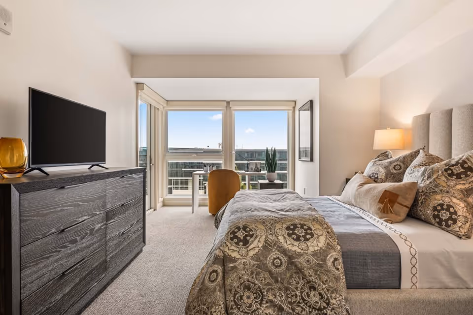 Well-lit bedroom with an upholstered bed and patterned bedding, a dresser with a TV, and floor-to-ceiling windows opening to a small balcony.